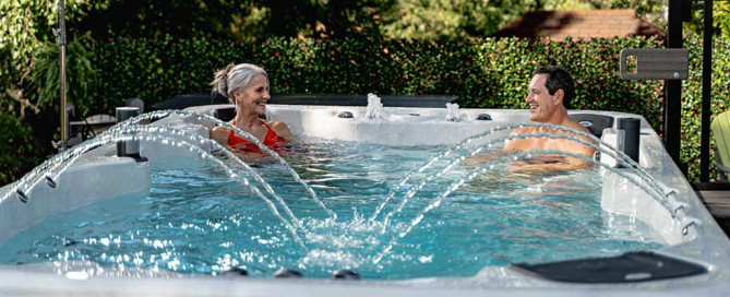Happy man and woman sitting opposite in corners of a large hot tub with crystal clear water and from sides a group of sprays come out of the spa. Hot tub is set in a garden with lots of shrubs and trees behind it. The Photo is of an Aspen Spa.