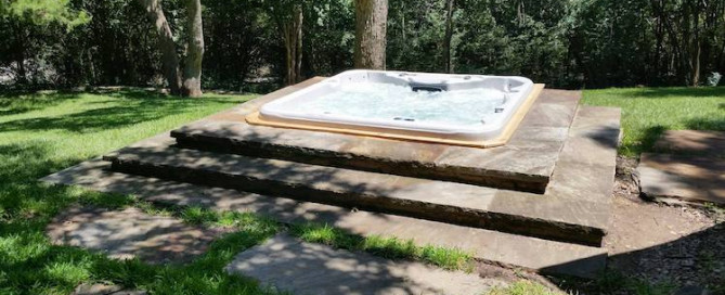 In wooded backyard's patio area, a 3-tiered stone platform surrounds a handsome hot tub set within it
