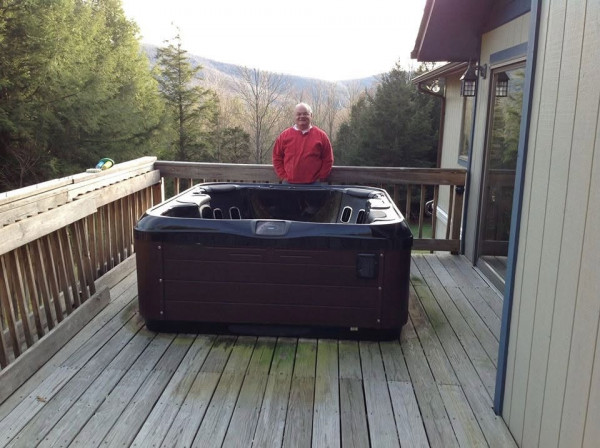 Happy looking customer standing behind his newly delivered hot tub; spa is sitting on his raised deck which is surrounded by large growth trees and in the background is a beautiful Mountain View